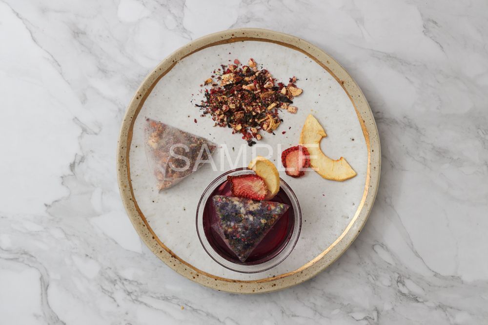 A tea sachet, a pile of loose herbal tea, and dried fruit slices arranged on a plate with a small bowl of brewed tea.