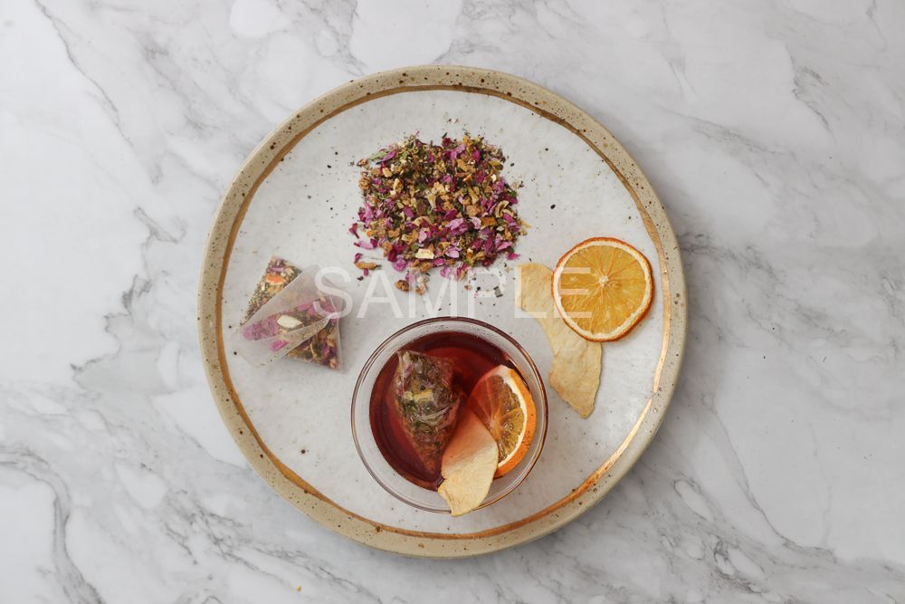 A top-down view of a ceramic plate with loose tea, tea bags, a cup of steeped tea with orange slices, and chips on marble.