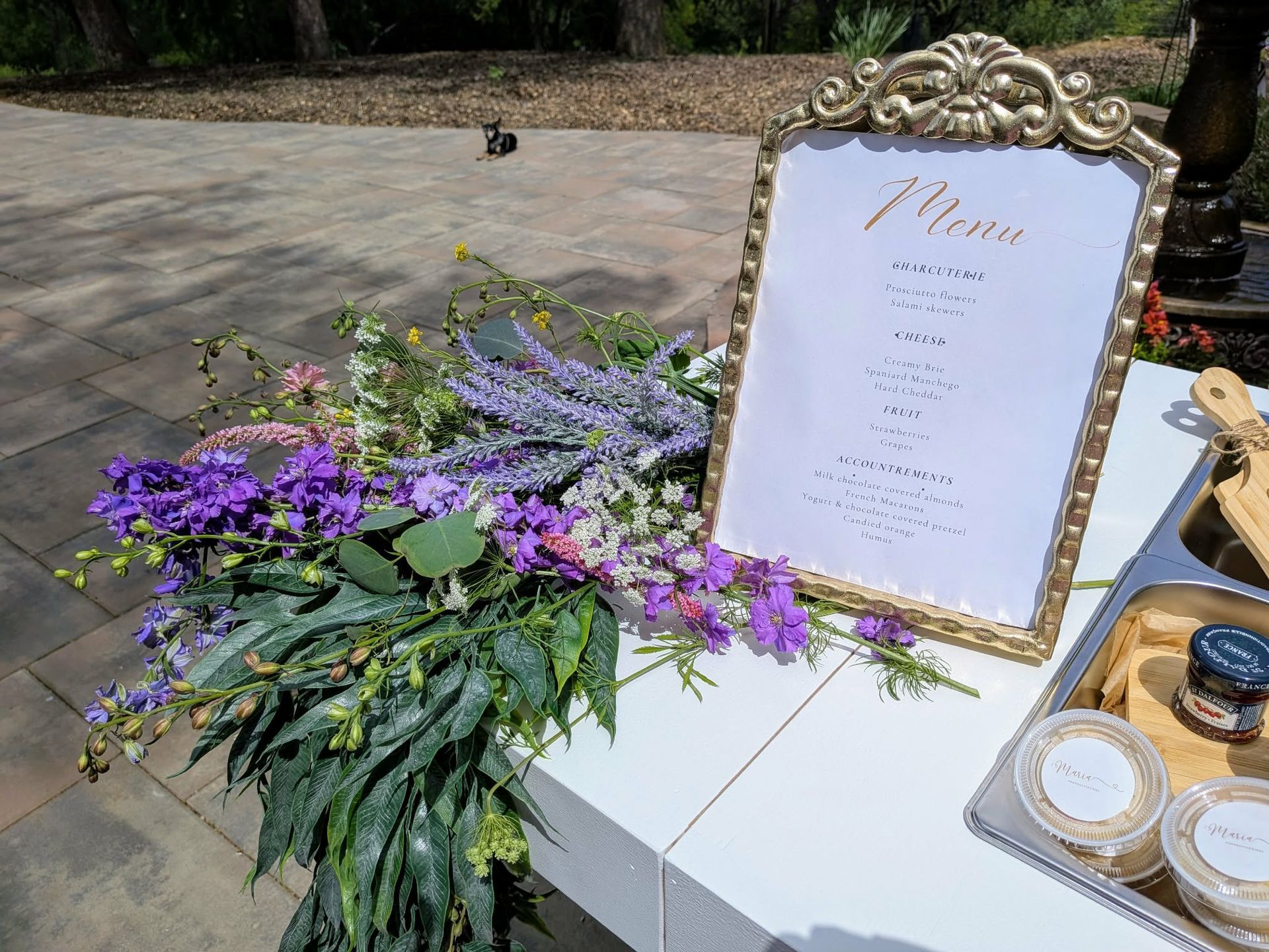 A menu board with gold frame and floral arrangement, outdoors. White table with food trays.