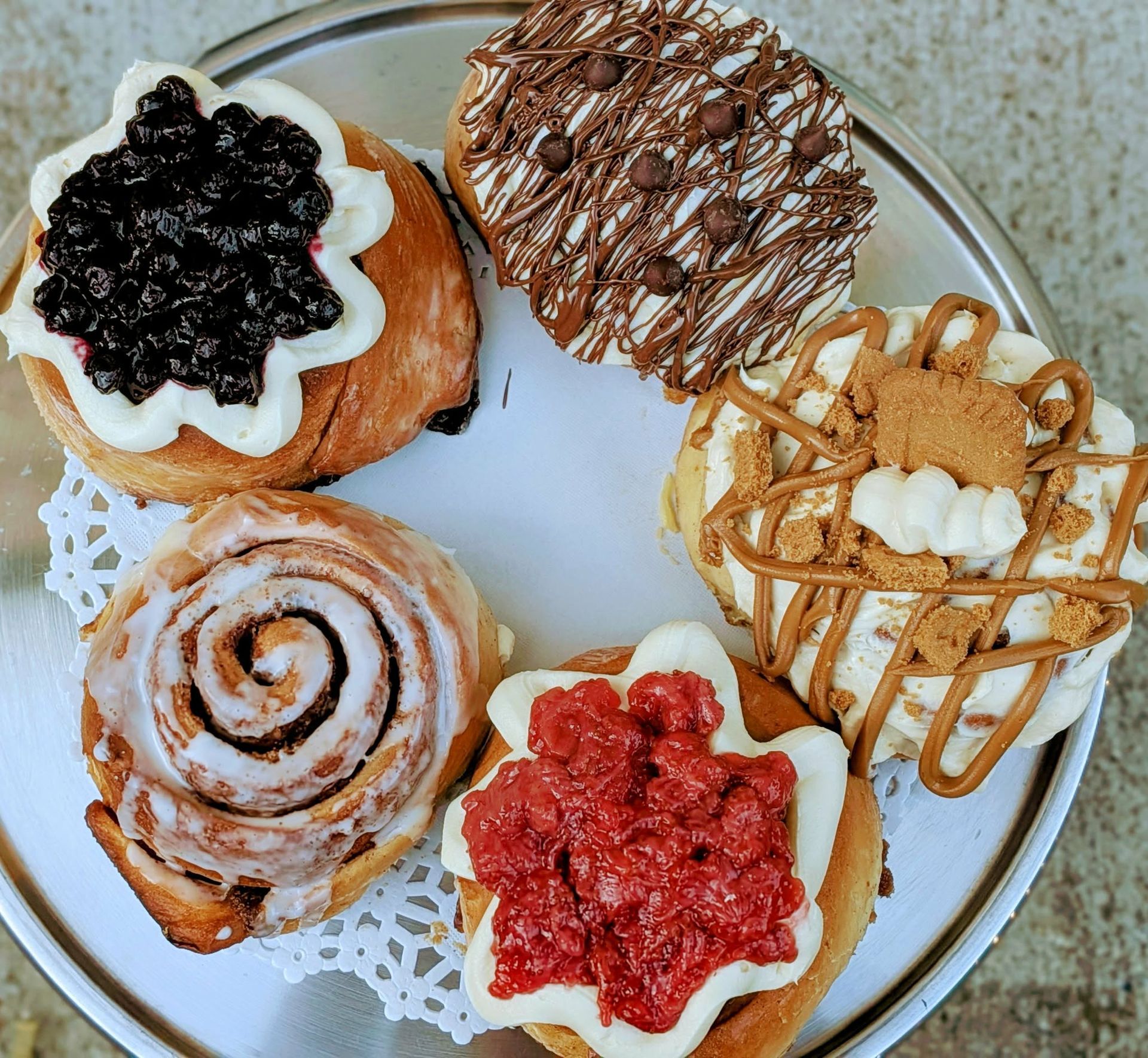 Assortment of decorated cinnamon rolls on a silver tray, featuring blueberry, chocolate, and other toppings.