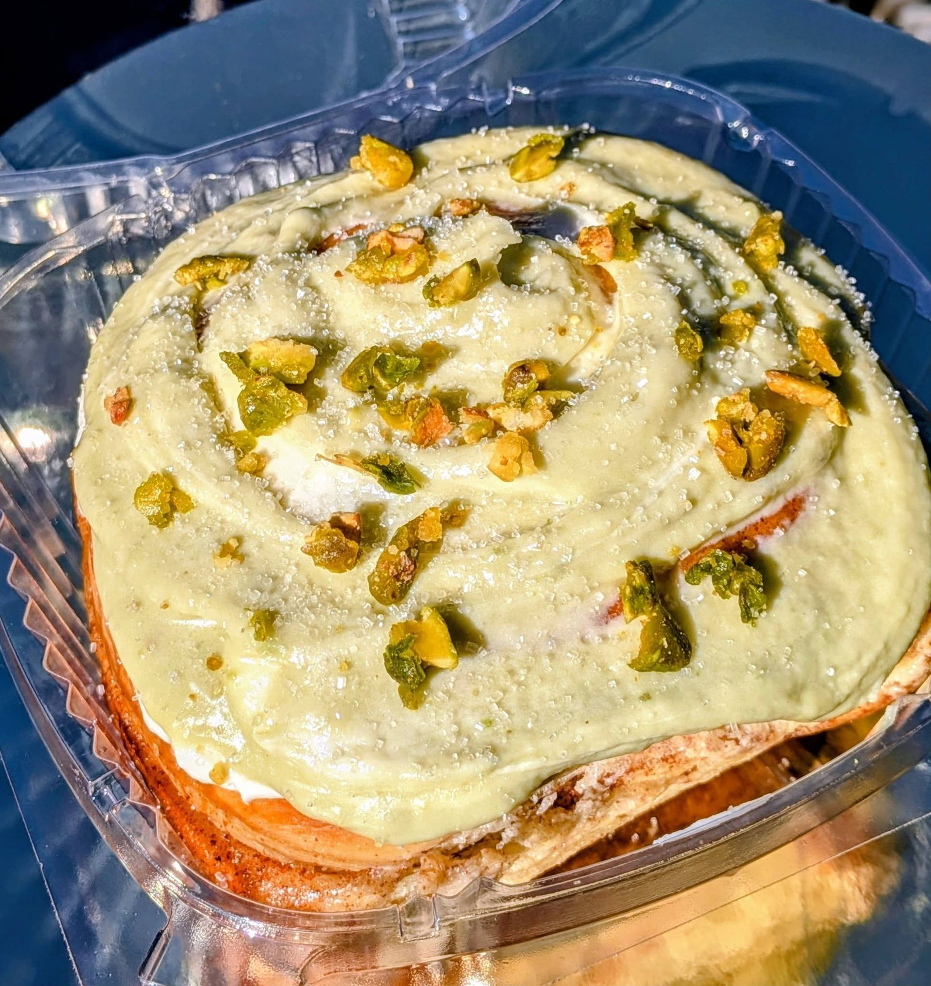 Pastry roll topped with pistachio cream and chopped pistachios in a clear container.