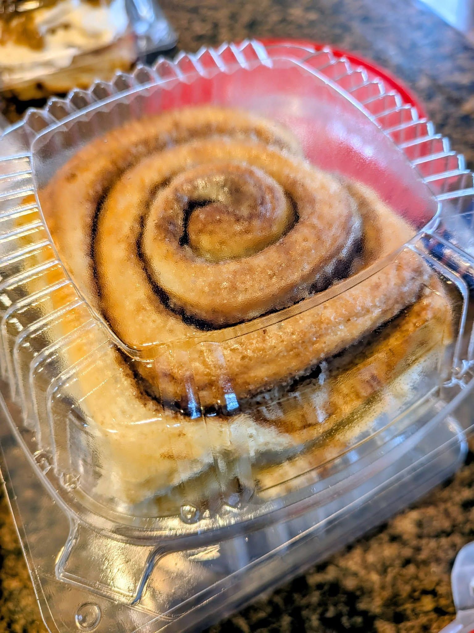 Cinnamon roll in clear plastic container, frosted with glaze.