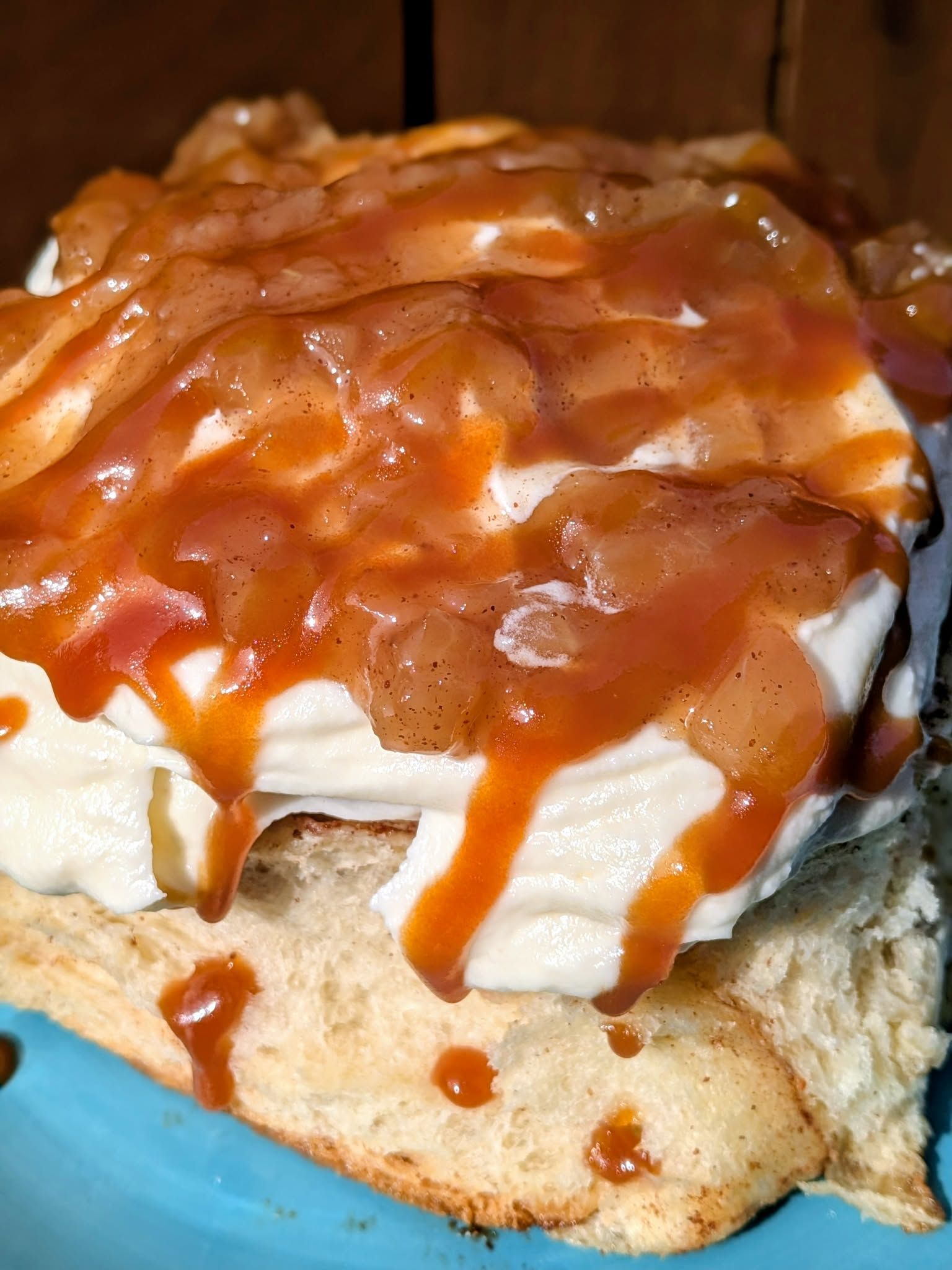 Cinnamon roll with thick cream topping, topped with apple pie filling and caramel sauce, on a blue plate.
