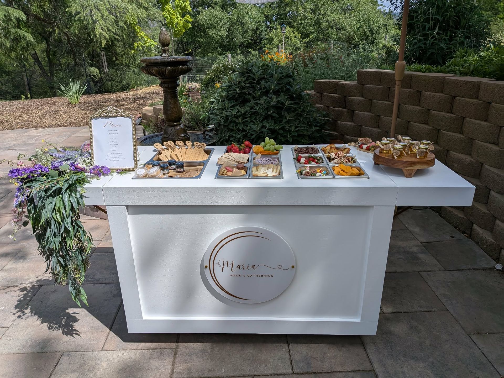 White food bar with various snacks, outdoors. Fountain and greenery in background.
