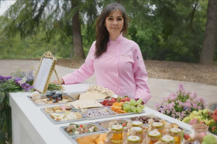 Woman in pink chef coat stands behind a buffet table with food outdoors.
