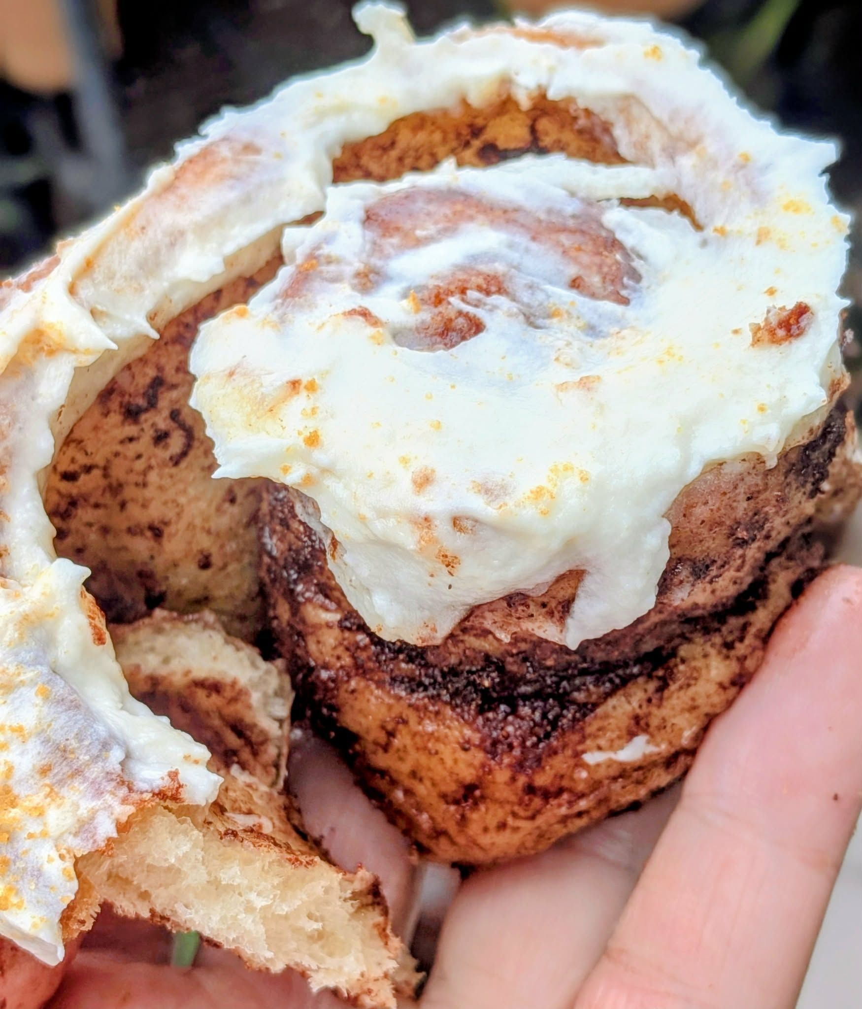 Close-up of a cinnamon roll with white icing, held in a hand. The roll is brown and fluffy, with visible layers.