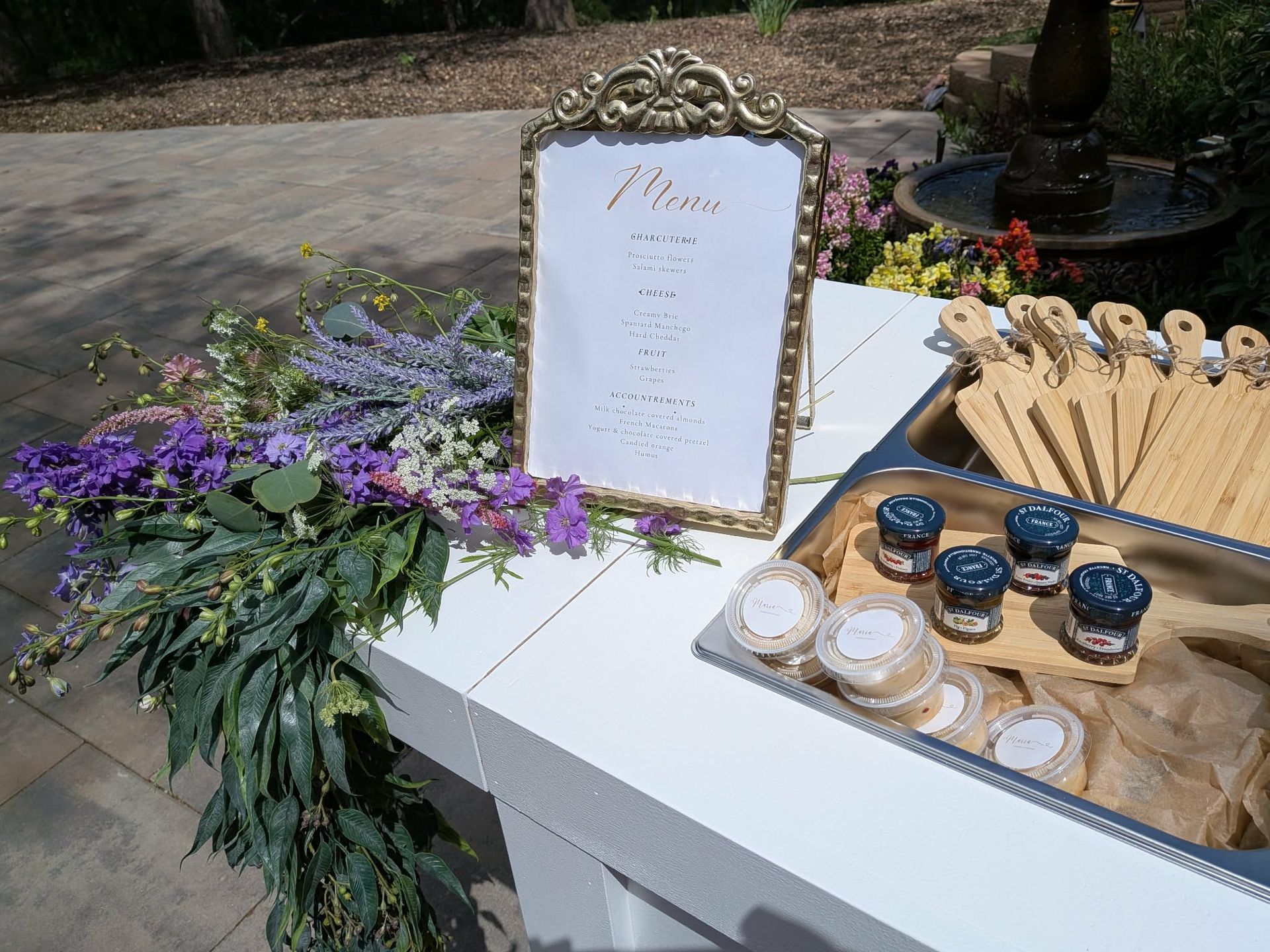 Menu board with gold frame, flowers, charcuterie setup, and fountain in background.
