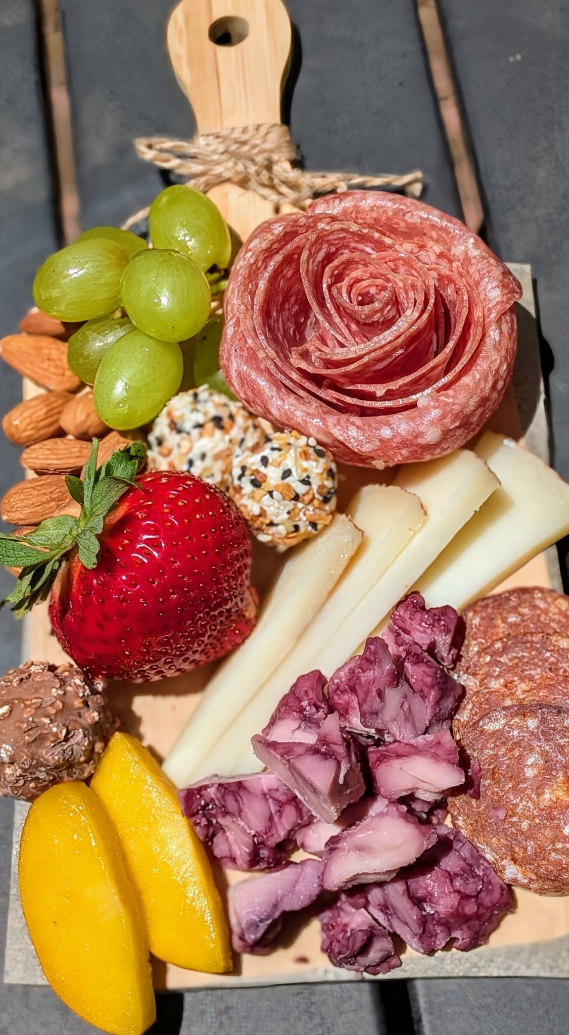 Charcuterie board: salami rose, cheese, grapes, strawberries, nuts, and chocolate on a wooden board.