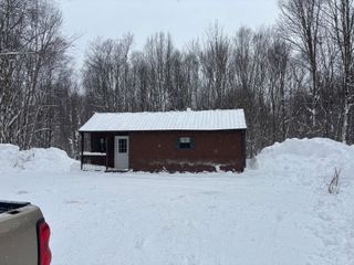 A small brown cabin with a metal roof surrounded by deep snow and a leafless forest in winter.