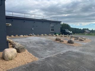 A gray building with a rooftop railing stands by an asphalt parking area bordered by decorative landscape stones.