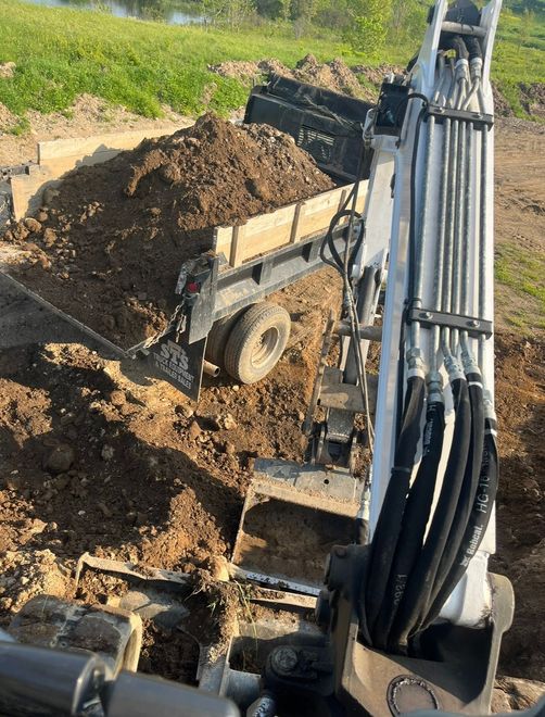 Excavator arm loading a pile of dark soil into a dump trailer on an outdoor site.