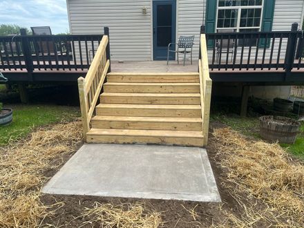 Wooden stairs lead up to a deck attached to a house, with a fresh concrete landing pad at the bottom on dirt and mulch.