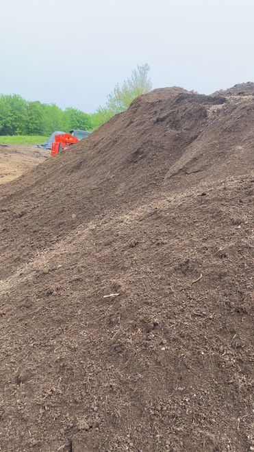 A large mound of dark soil or mulch at a construction site with an orange piece of machinery visible in the background.