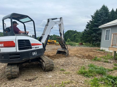 A person operates a white Bobcat E72 excavator to dig dirt in a yard next to a gray house.