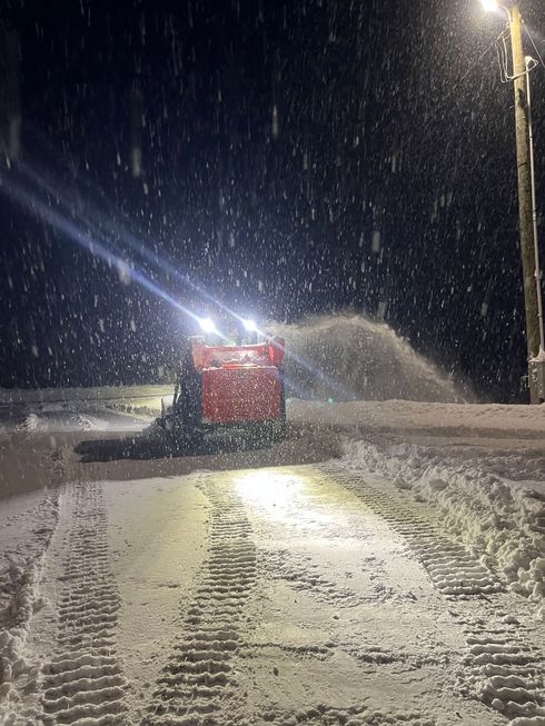 A red snowblower clears snow at night, with bright lights illuminating the falling flakes and tracked tread marks.