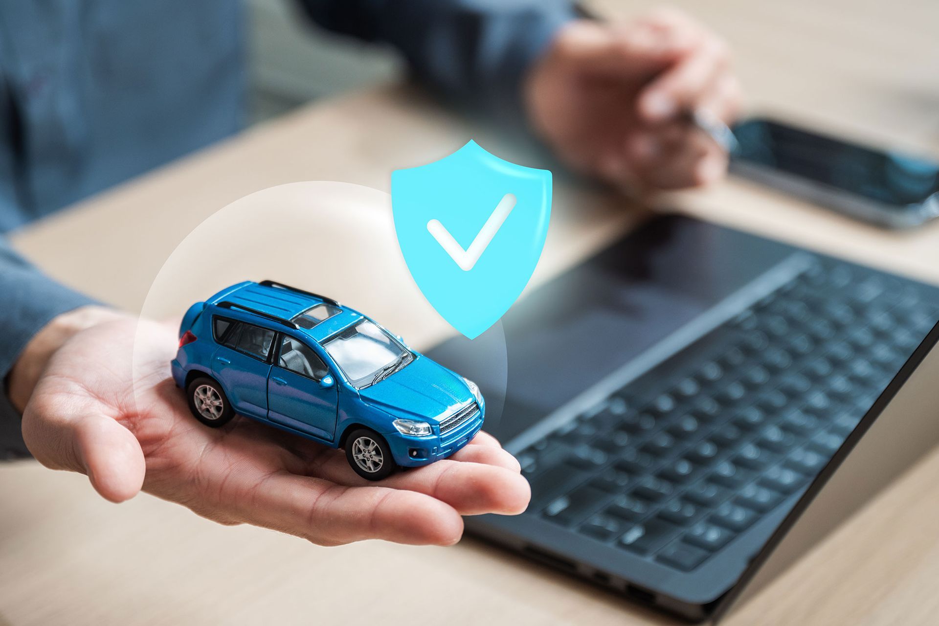Person's hand holding a blue toy car with a shield icon above, next to a laptop and phone.