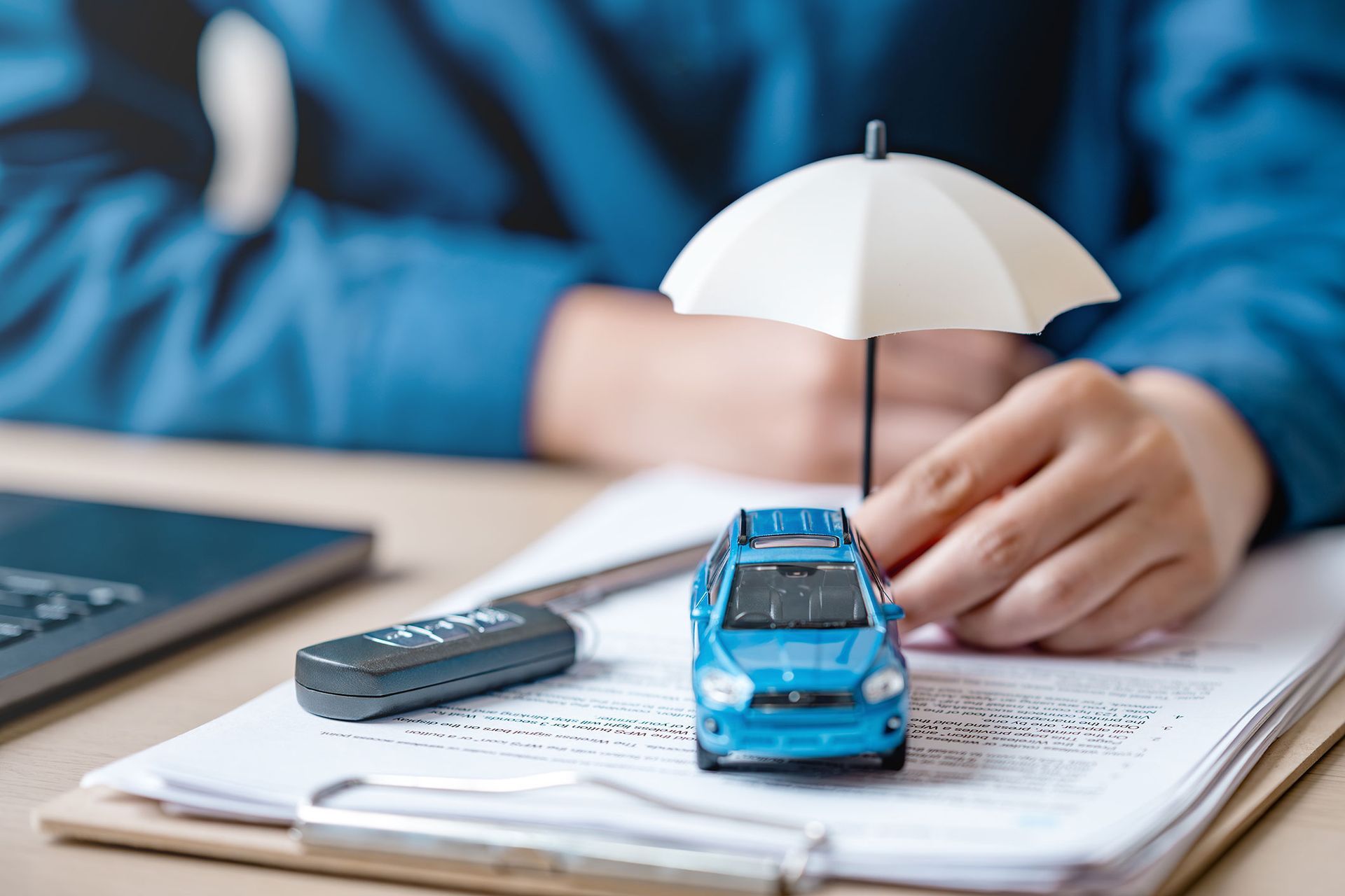 Blue toy car under a small umbrella on paperwork, with a remote and laptop.