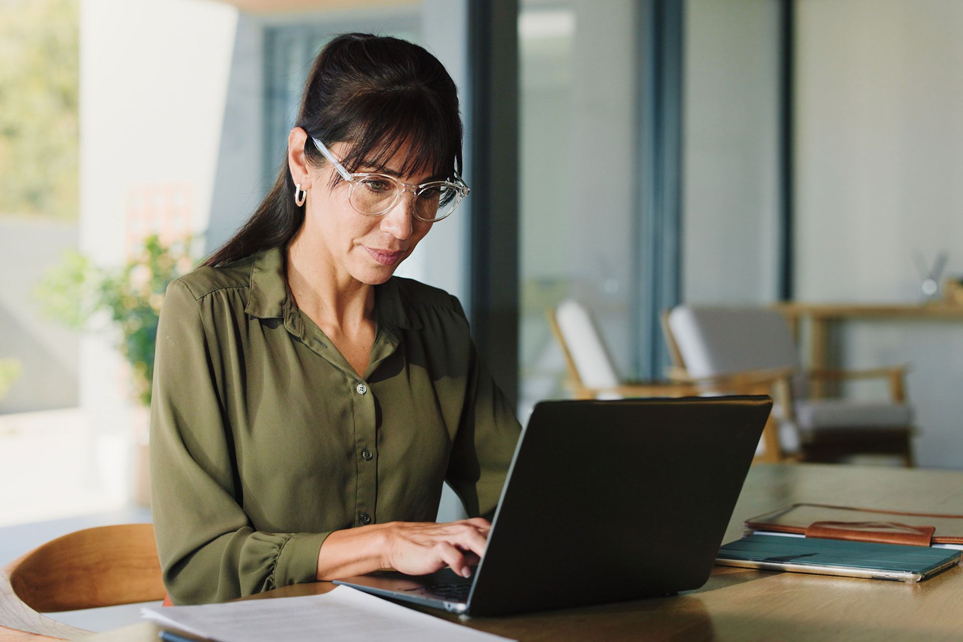 Woman wearing glasses typing on a laptop at a table. Olive green shirt, sunny setting.