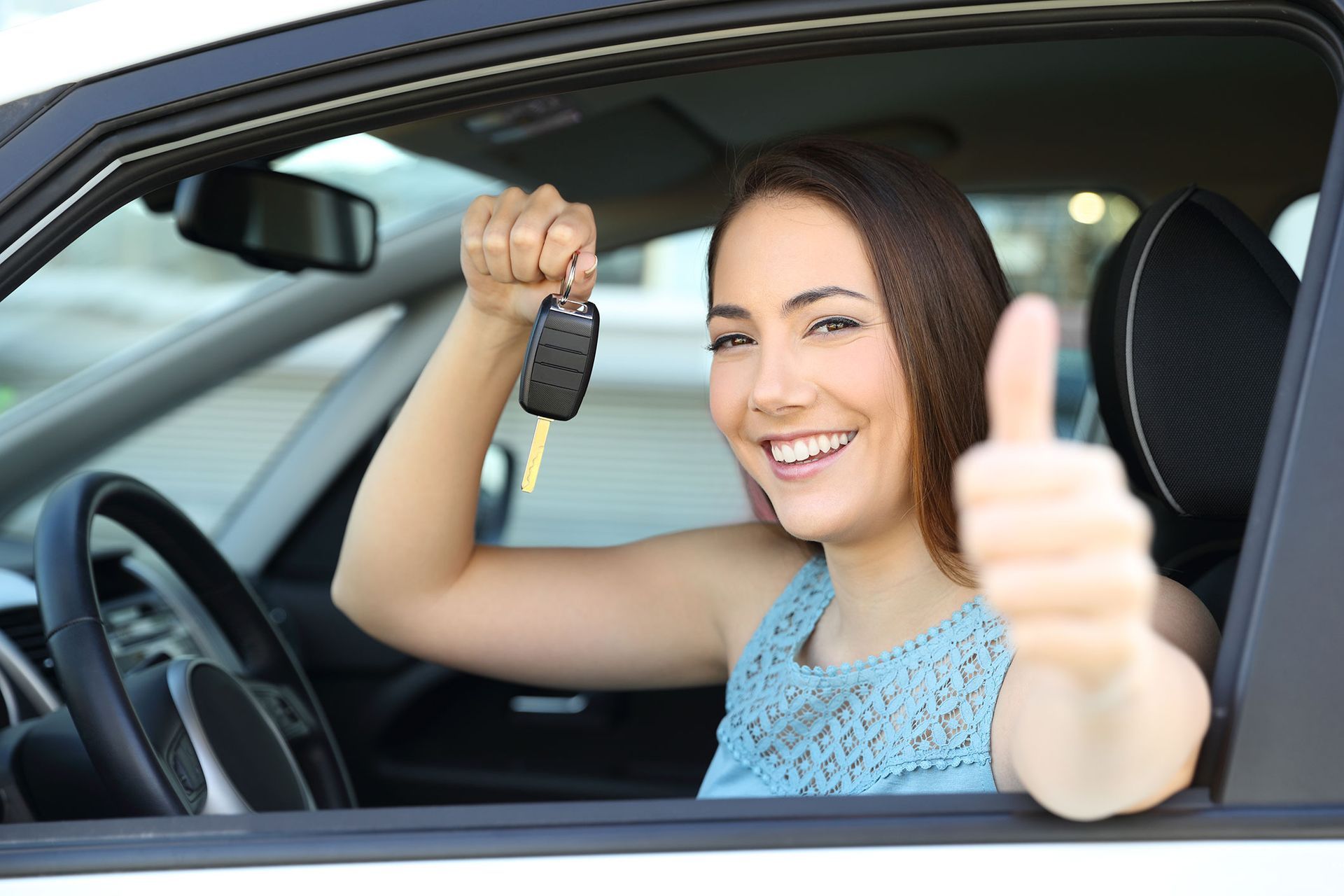 Woman in car holding keys, smiling, giving thumbs up.