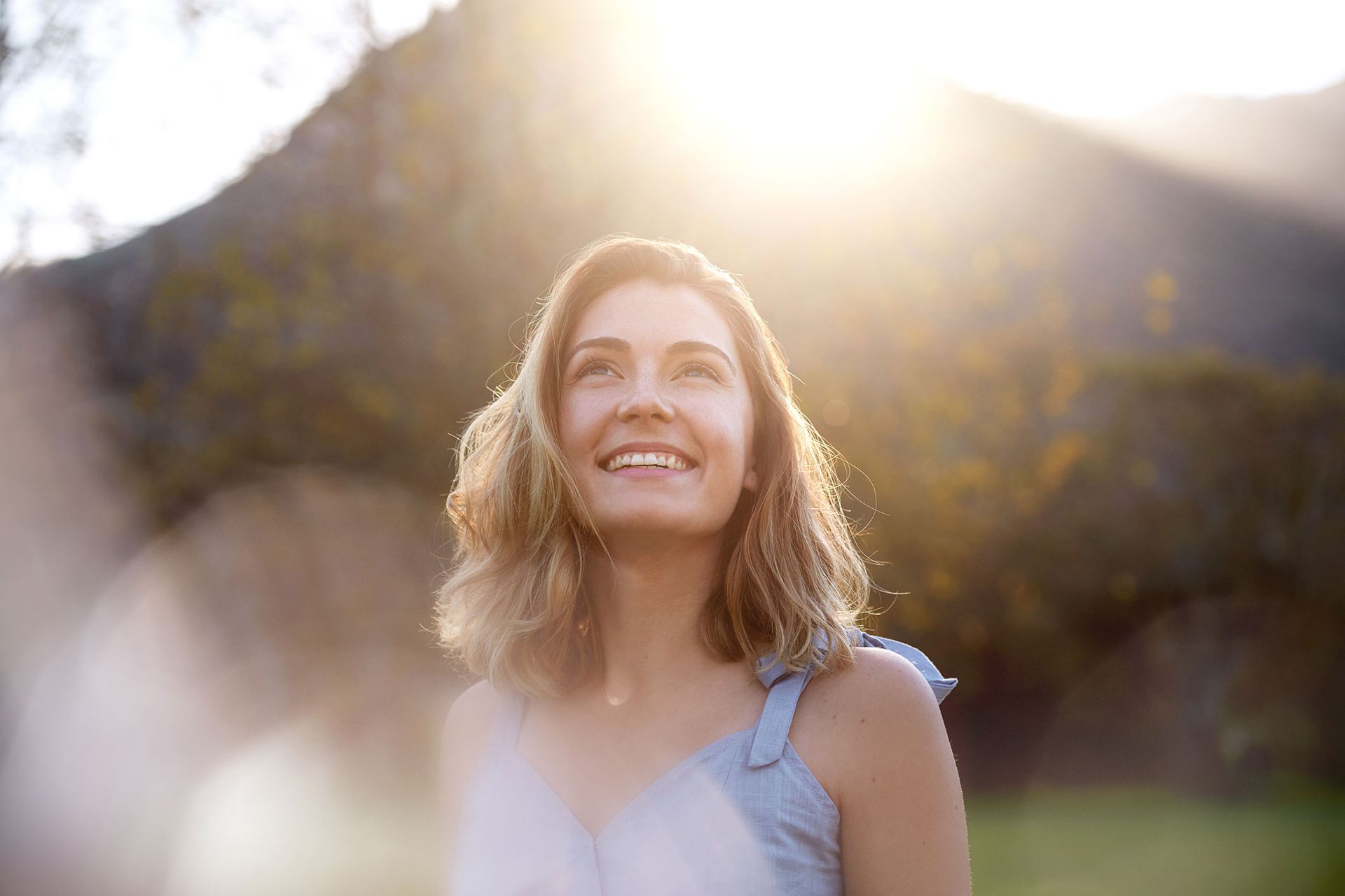 Woman looking upward, smiling, bathed in sunlight. Background: trees, mountains.