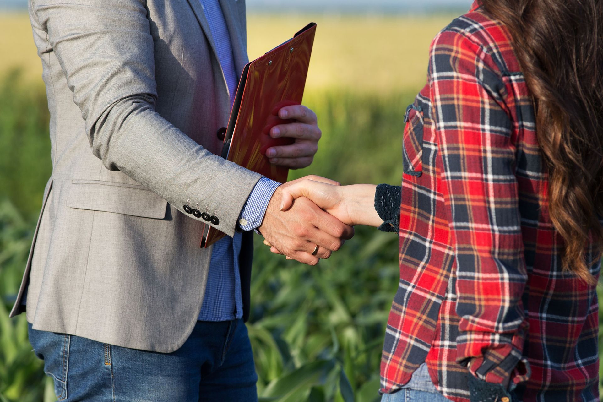 Man in suit shakes hands with person in red plaid shirt; field in background.