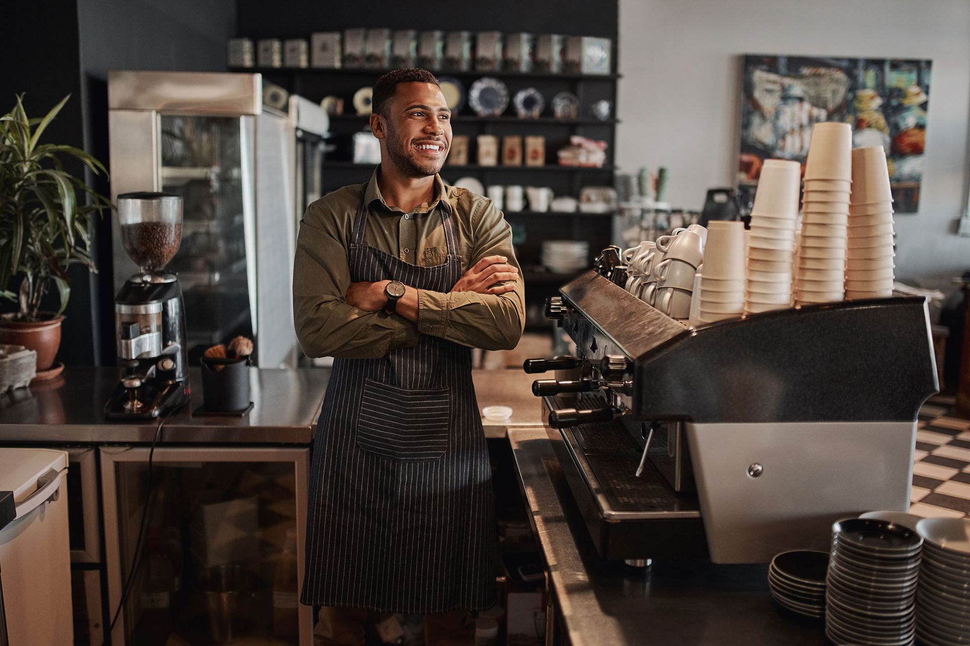 Barista smiles in his coffee shop with arms crossed; espresso machine and cups visible.