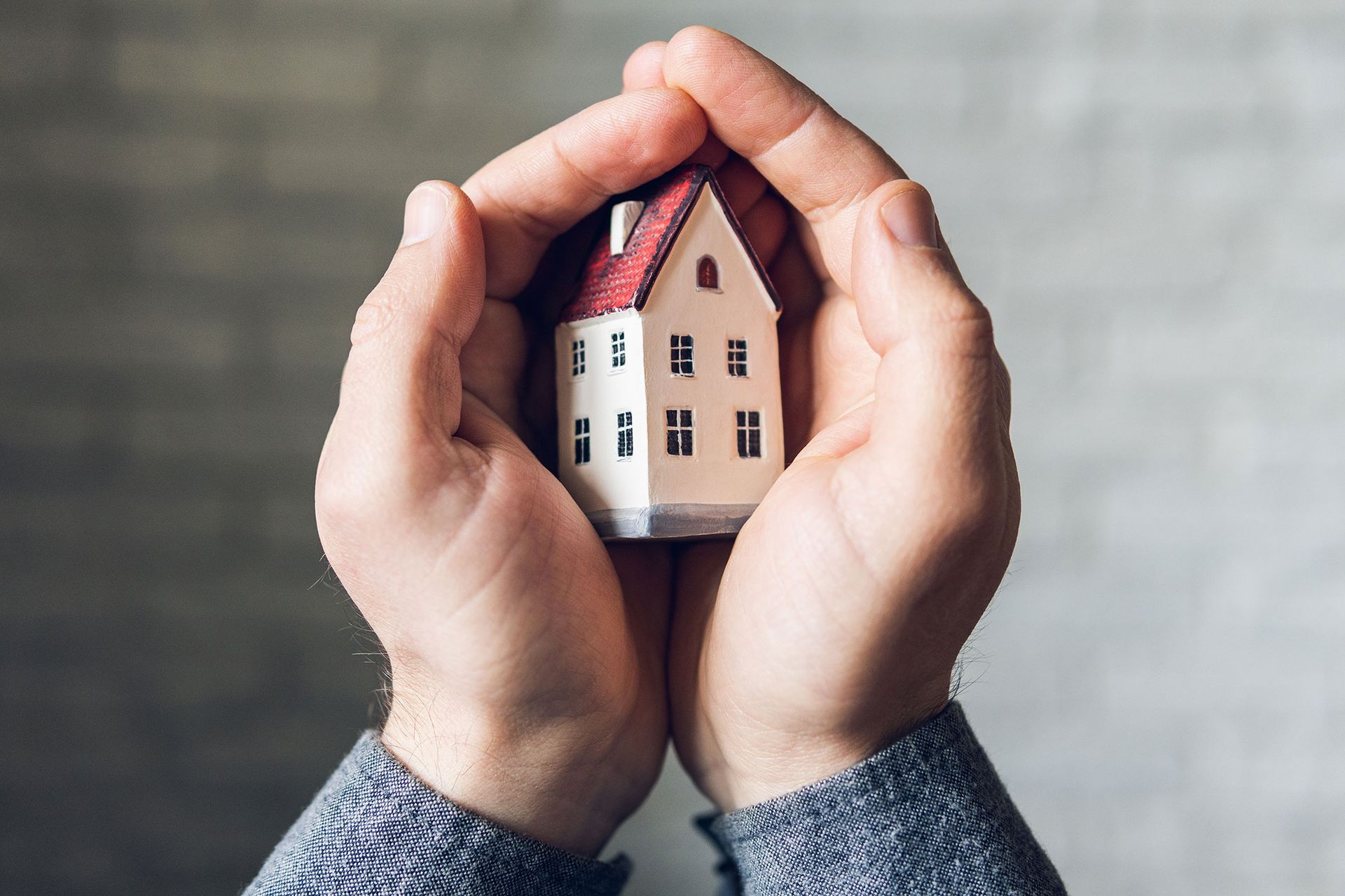 Hands cupping a small model house with a red roof, against a gray background.