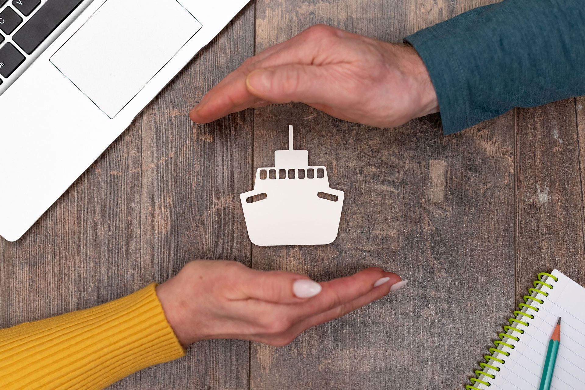 Hands cupped around a cutout ship, signifying insurance or protection, on a wooden surface with a laptop and notepad.