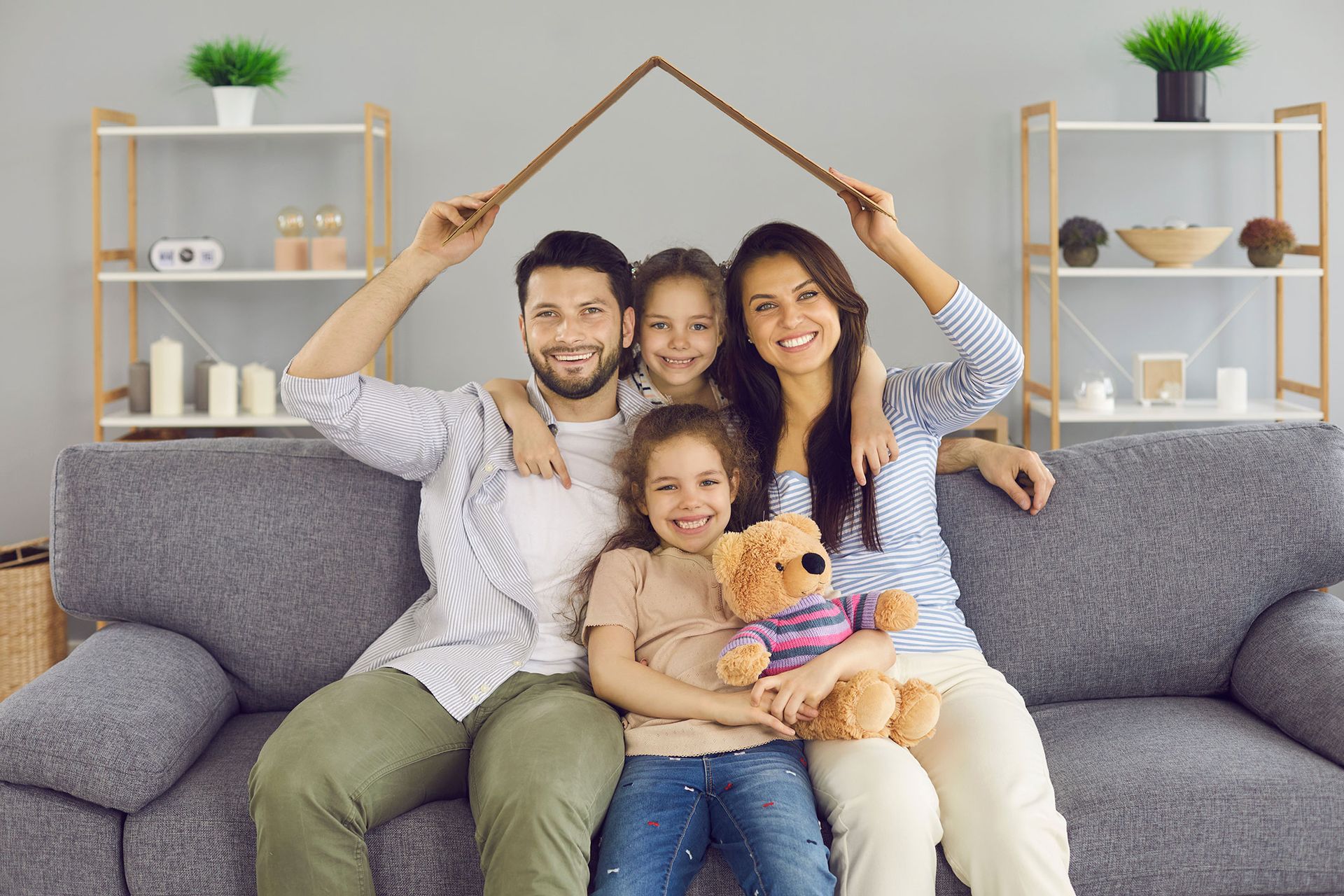 Family on couch, holding house-shaped structure above them. Smiling, in a living room.