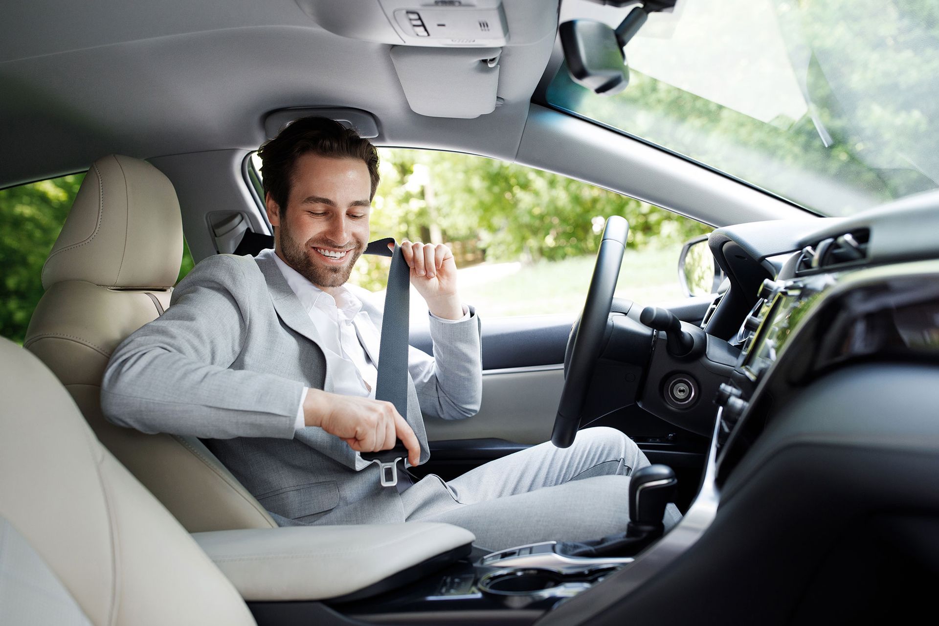 Man in a gray suit smiles while fastening his seatbelt inside a car.