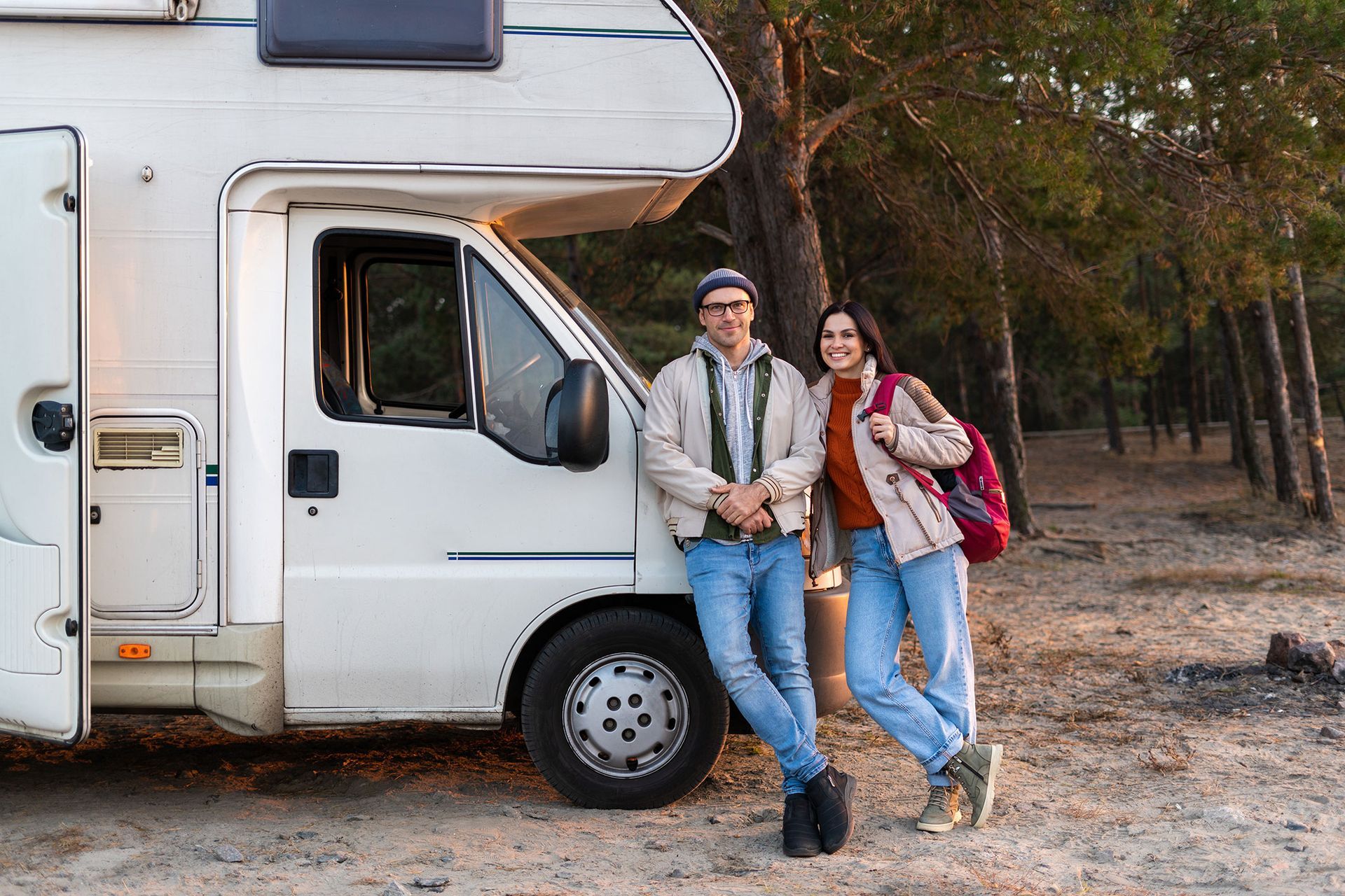 Couple posing by a white RV in a wooded area, smiling.