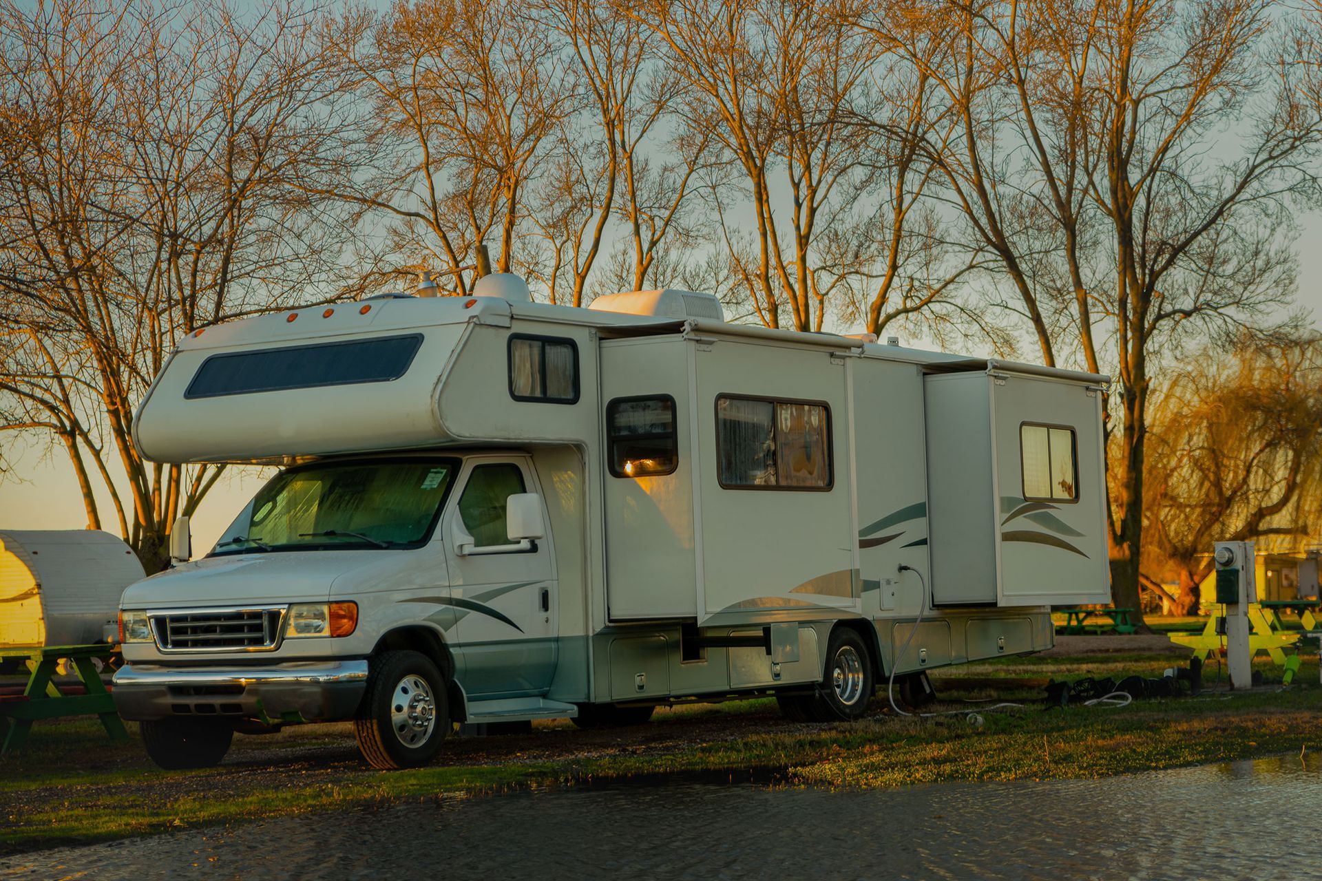 White RV parked in a campground, trees in the background, awning extended, evening sunlight.