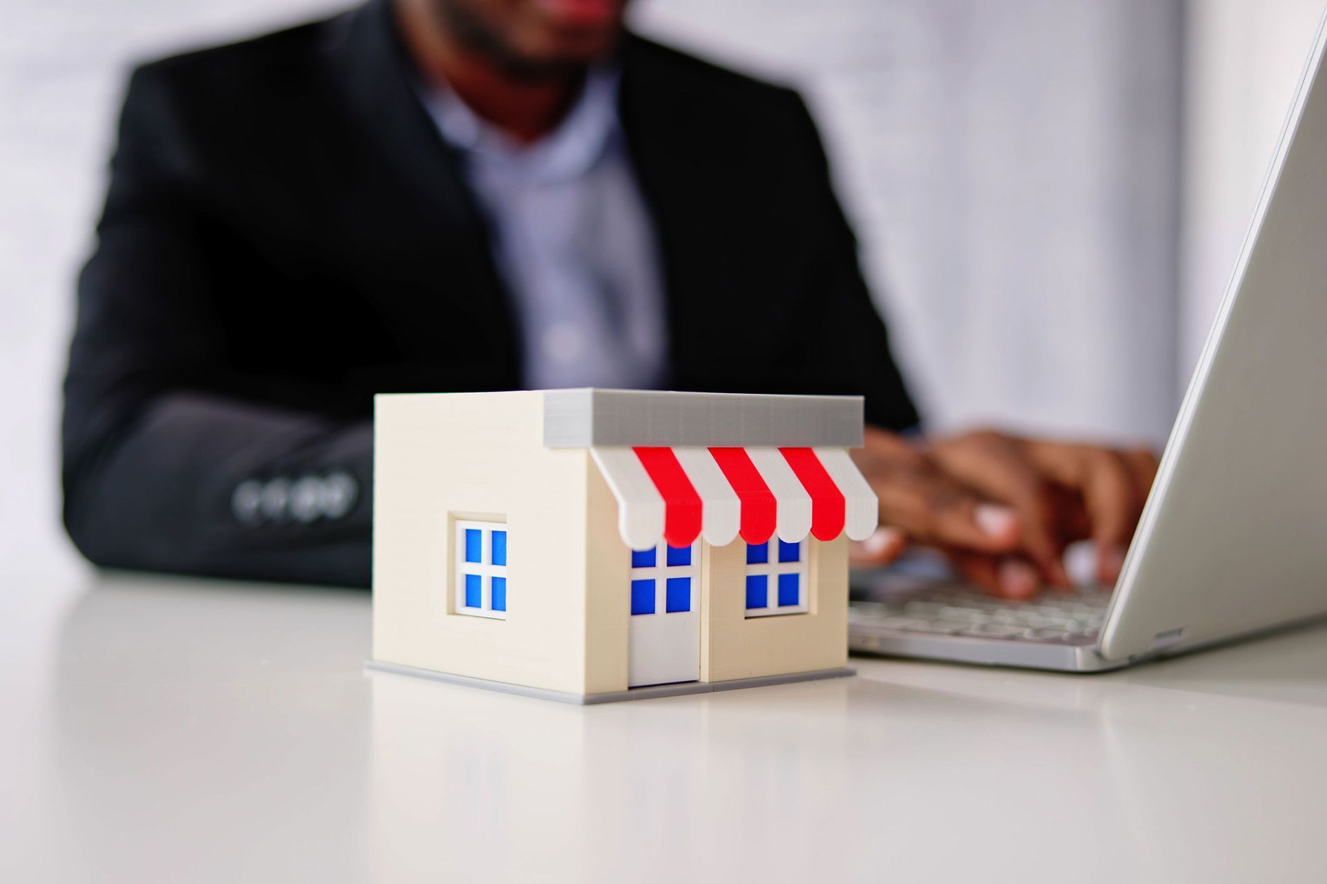A person in a suit works on a laptop next to a miniature store model with a red and white awning.