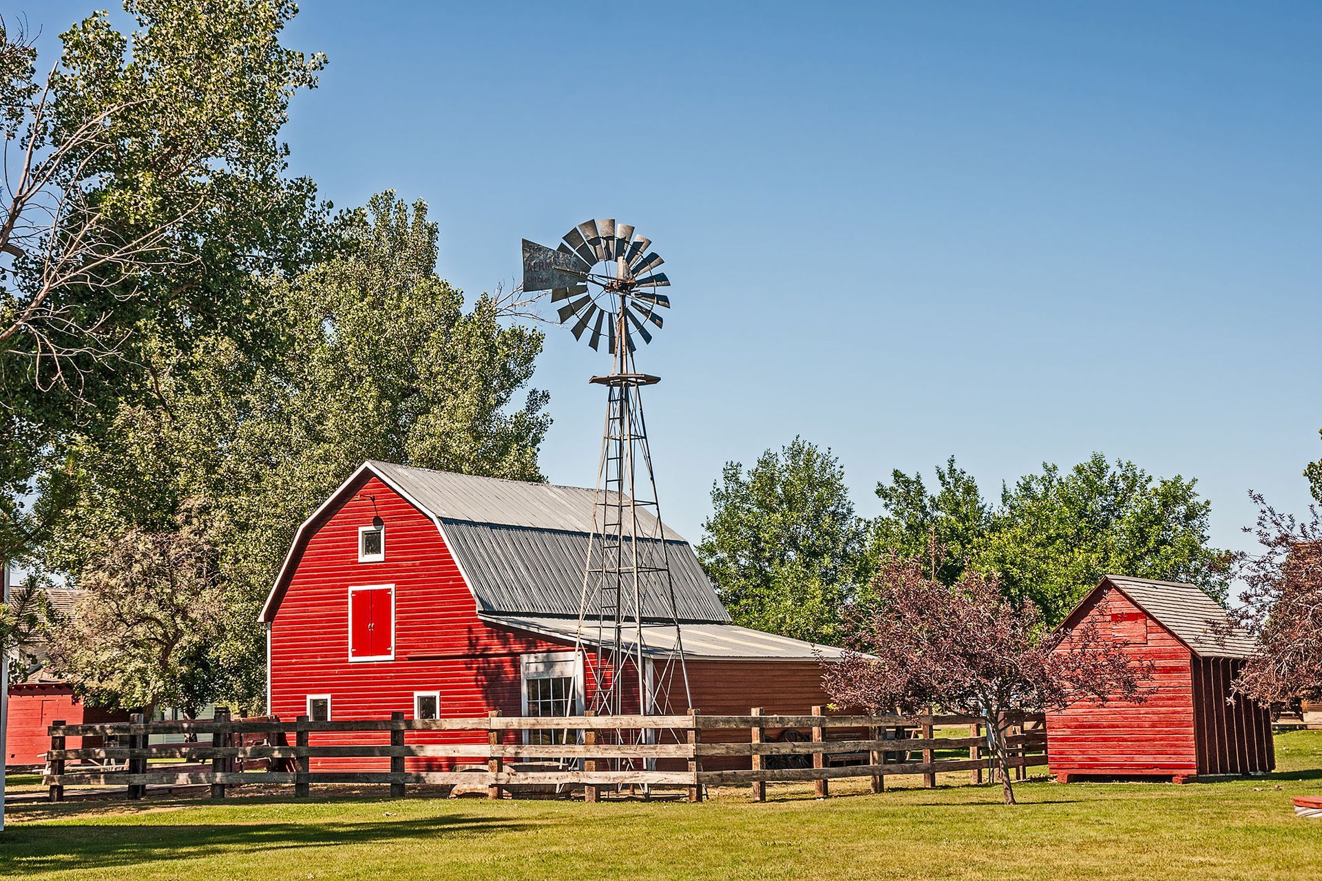 Red barn and shed with windmill on a grassy field, under a bright blue sky.