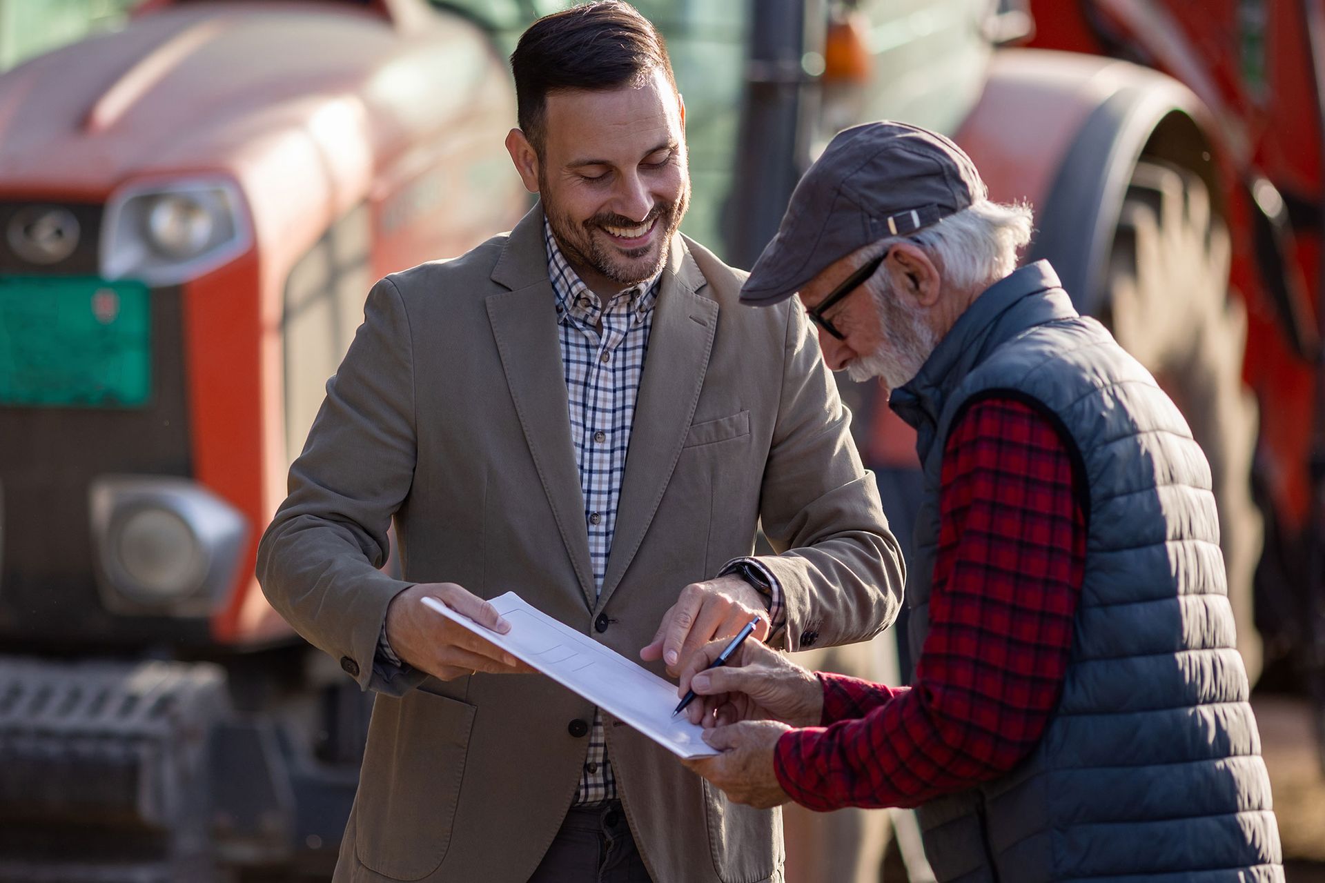 A man in a suit shows documents to a farmer in front of a tractor.