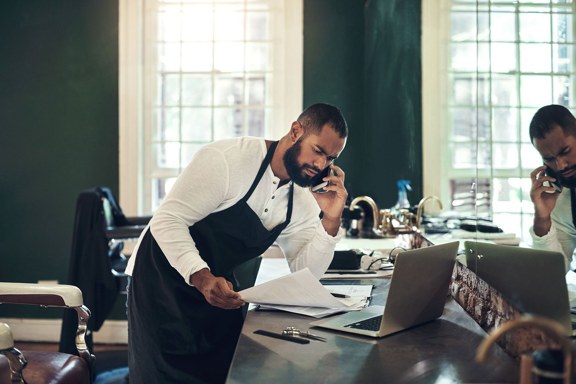 Two men, both in aprons, on phone calls at a desk with a laptop and documents in a barber shop.