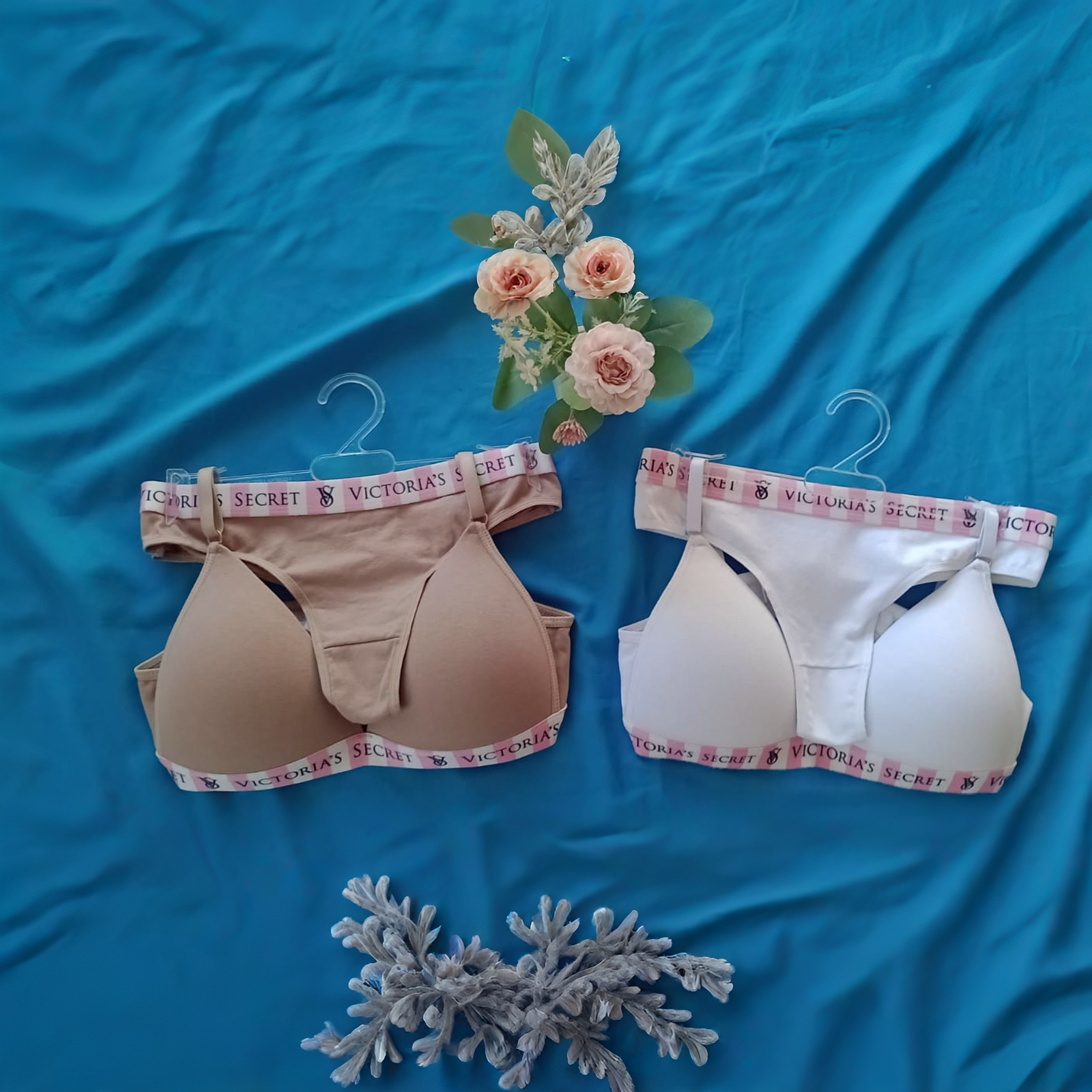 Dos sujetadores con bandas elásticas de marca, uno beige y otro blanco, sobre un fondo azul decorado con flores artificiales.