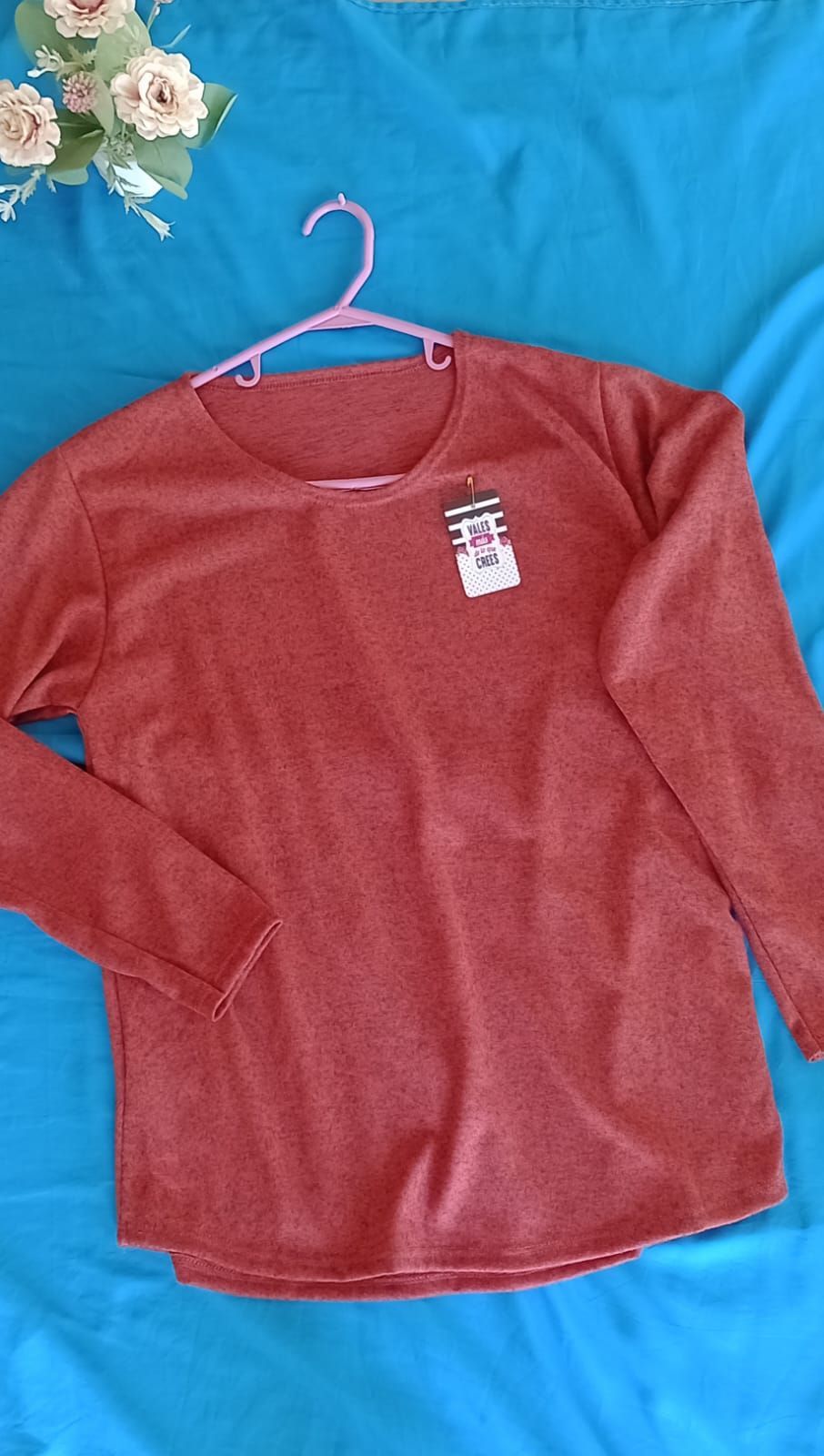 Una blusa de manga larga de color naranja óxido liso cuelga de una percha rosa sobre un fondo azul brillante.