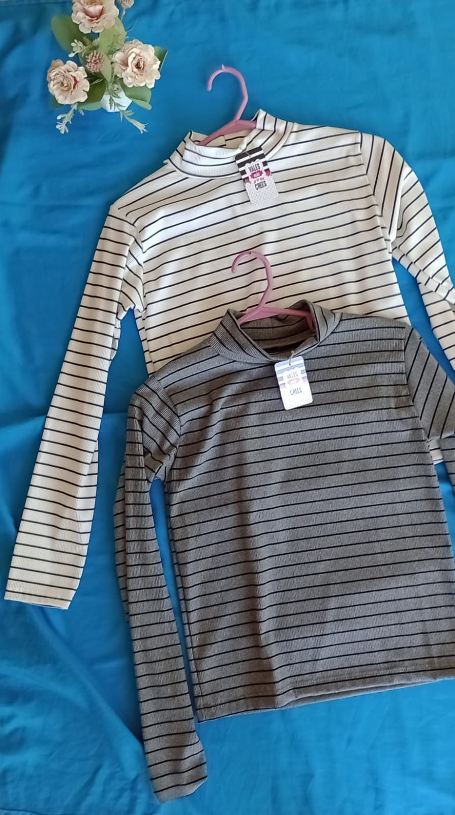 Una camisa blanca de manga larga con rayas negras horizontales colgada en una percha rosa sobre un fondo azul.