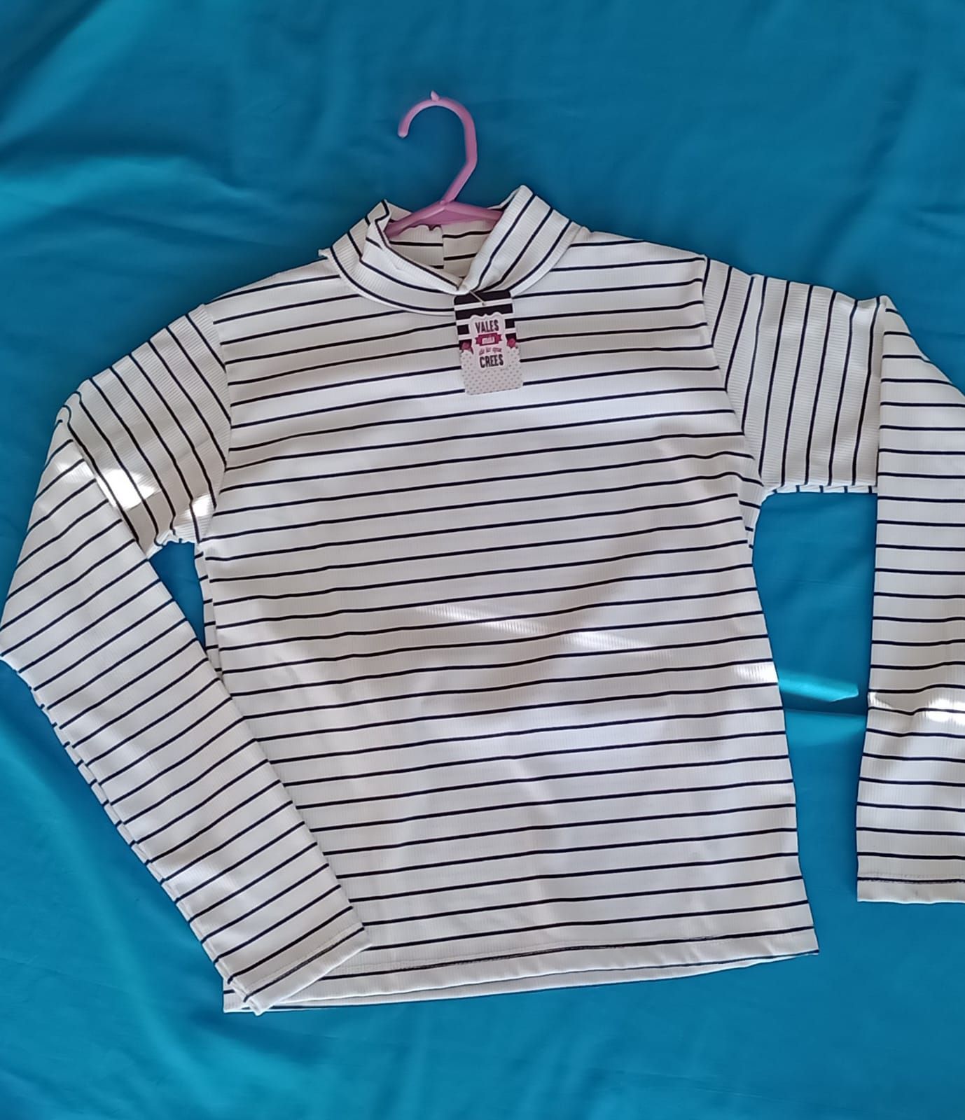 Una camisa blanca de manga larga con rayas negras horizontales colgada en una percha rosa sobre un fondo azul.