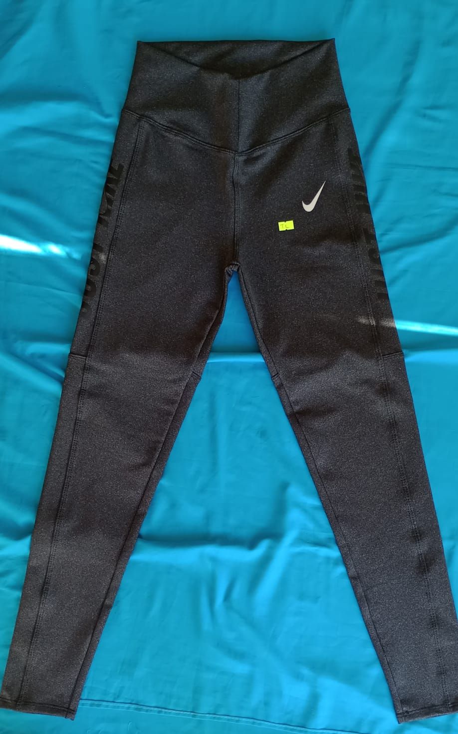 Leggings Nike de talle alto en color gris oscuro con el logotipo swoosh blanco, extendidos sobre una superficie azul.