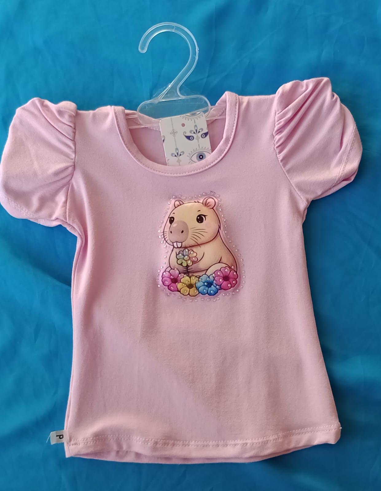 Una camisa rosa claro con mangas abullonadas, con un dibujo animado de un capibara sentado entre flores en el pecho.