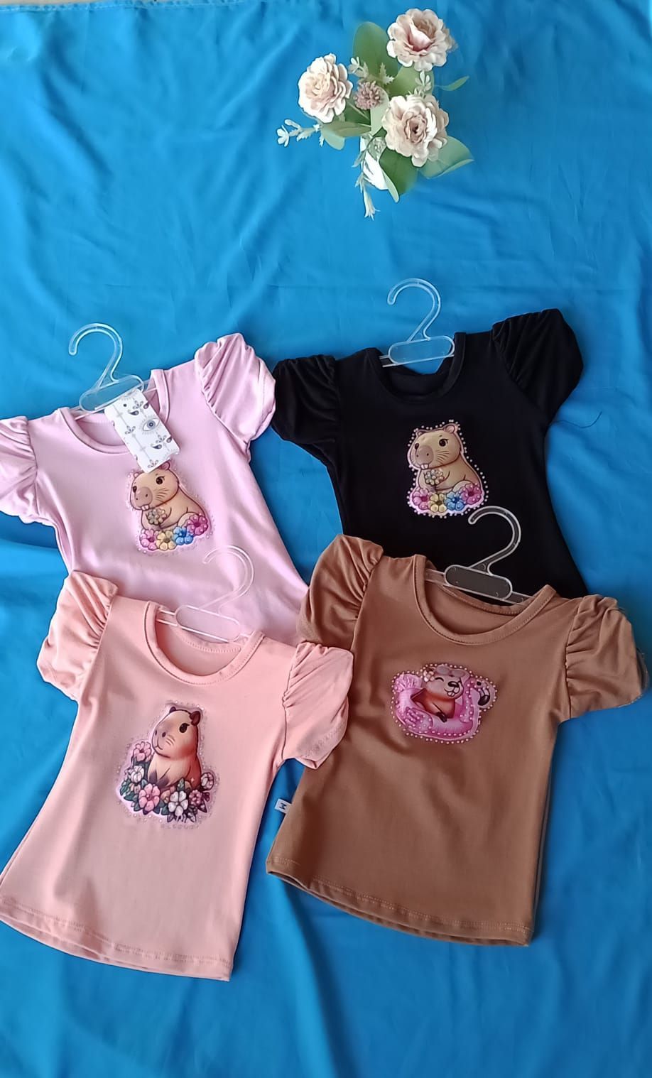 Una camisa rosa claro con mangas abullonadas, con un dibujo animado de un capibara sentado entre flores en el pecho.