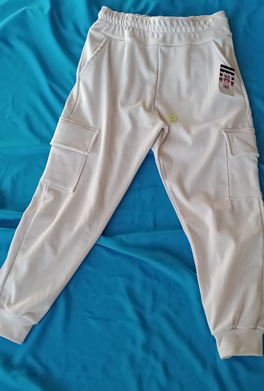 Pantalones cargo deportivos de color blanco roto con puños elásticos y bolsillos laterales sobre fondo azul.