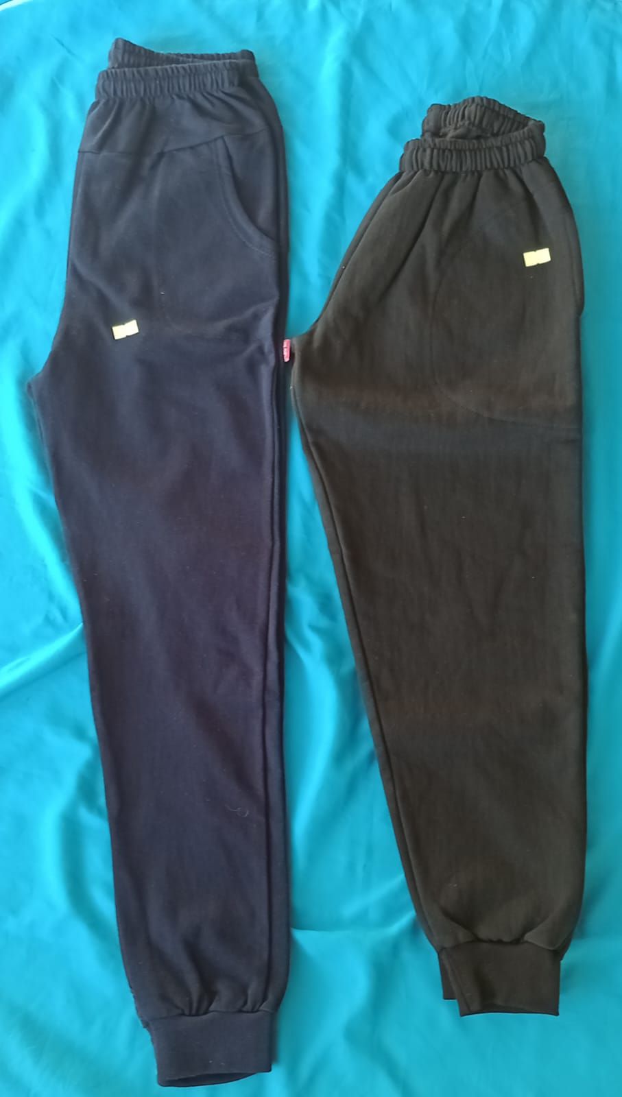 Dos pares de pantalones deportivos, uno azul marino y otro negro, extendidos sobre una superficie azul.