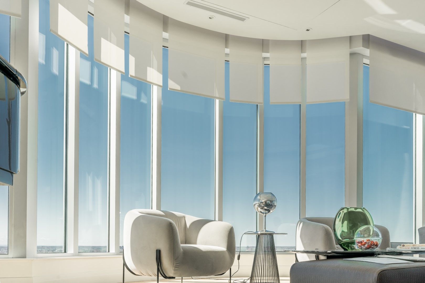 Bright, modern room with large windows, white furniture, and partially lowered blinds overlooking the sea.