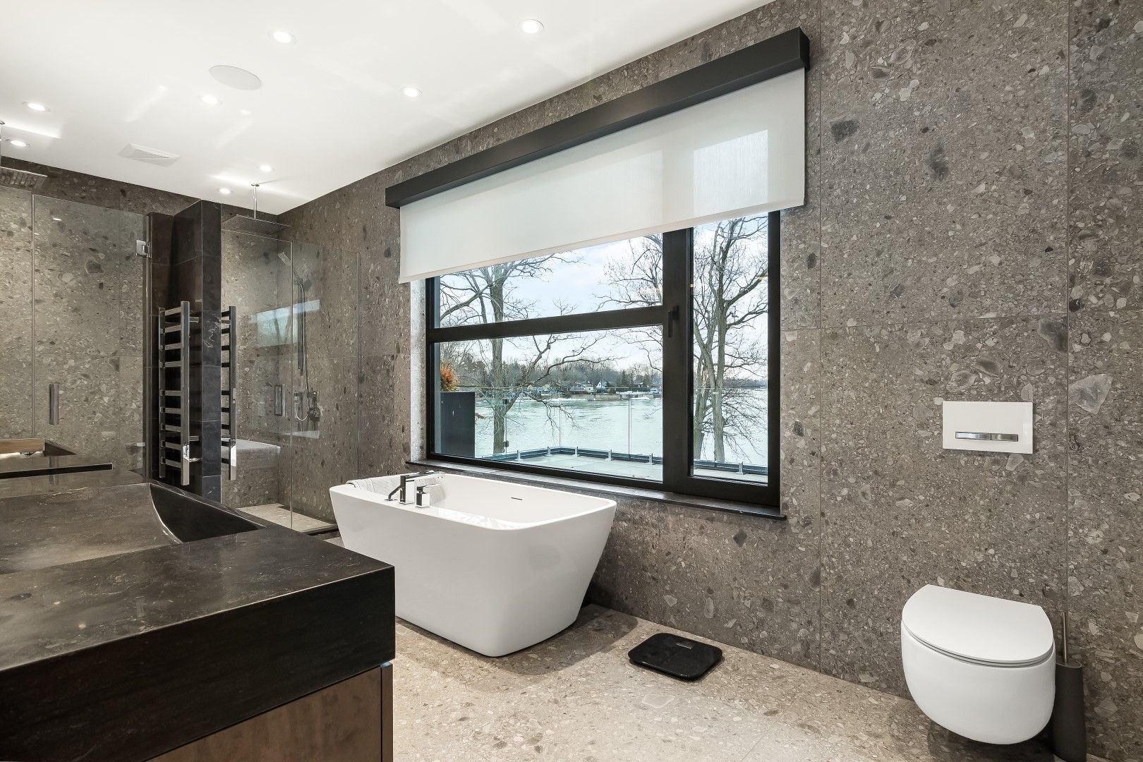 Modern bathroom with gray stone walls, white soaking tub, and large window overlooking a body of water.