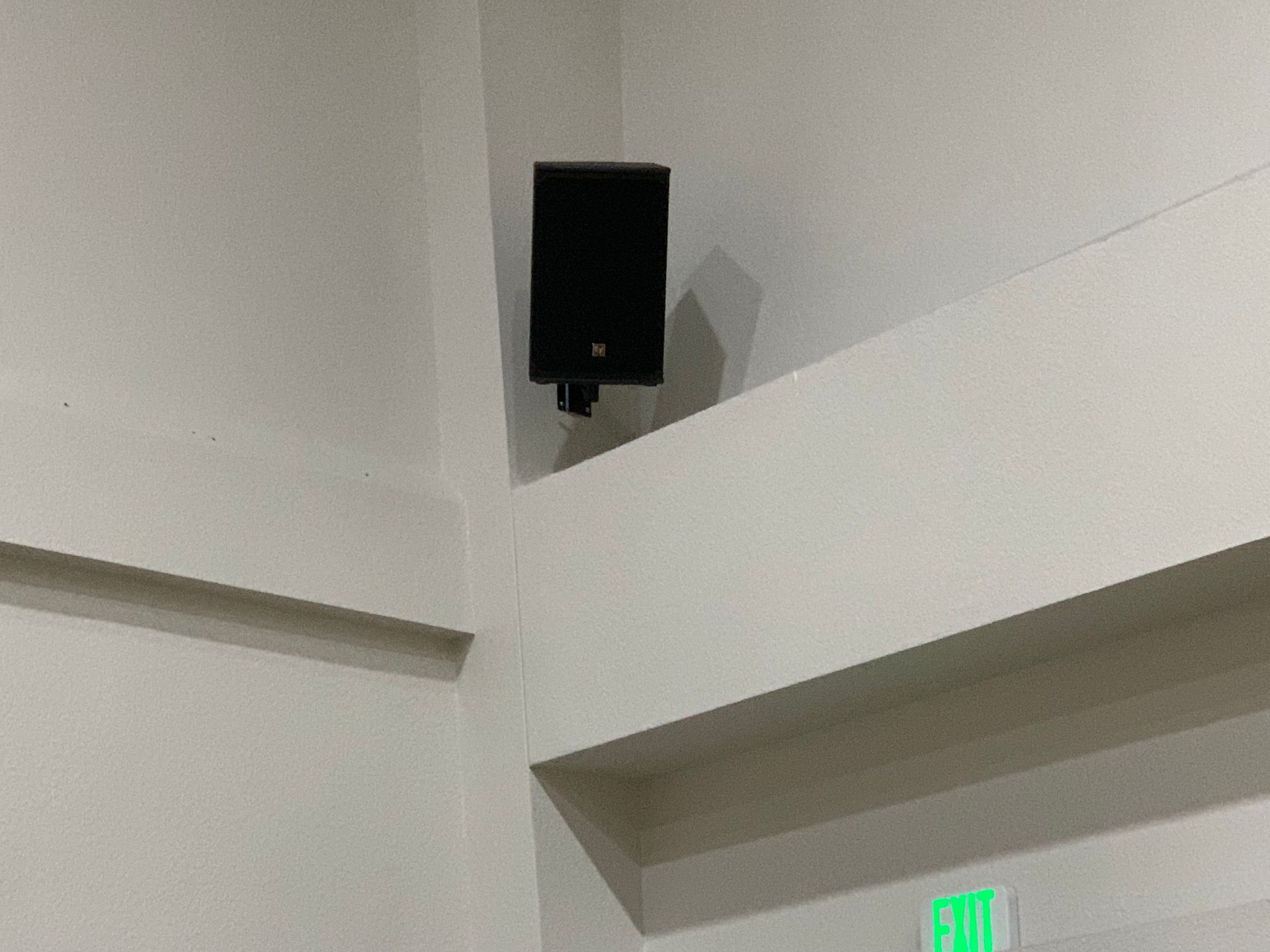 A speaker is hanging from the ceiling next to a green exit sign.