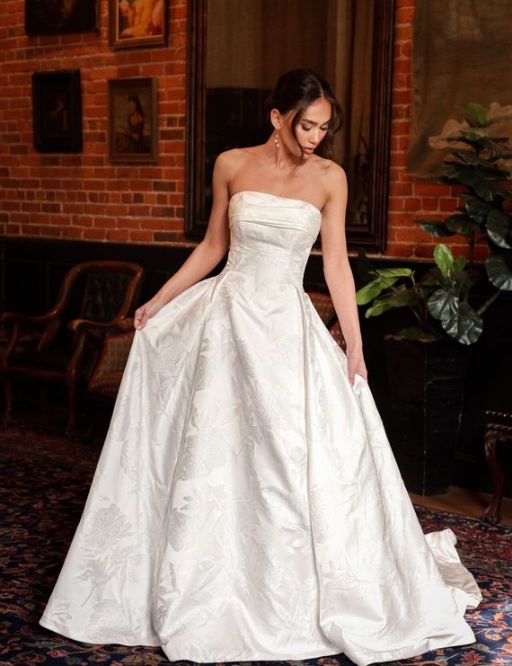 A woman in a white wedding dress is standing in front of a brick wall.
