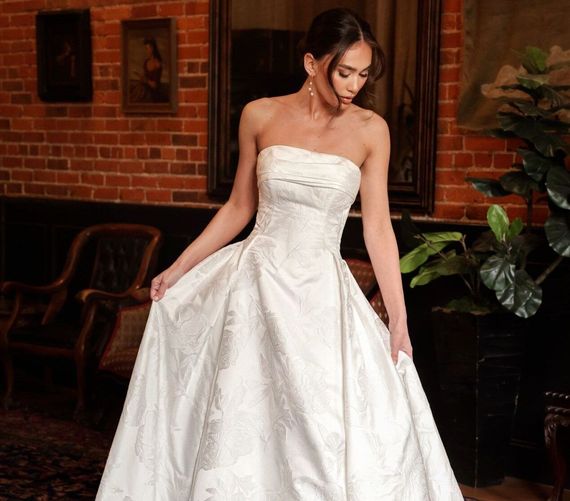 A woman in a white wedding dress is standing in front of a brick wall.